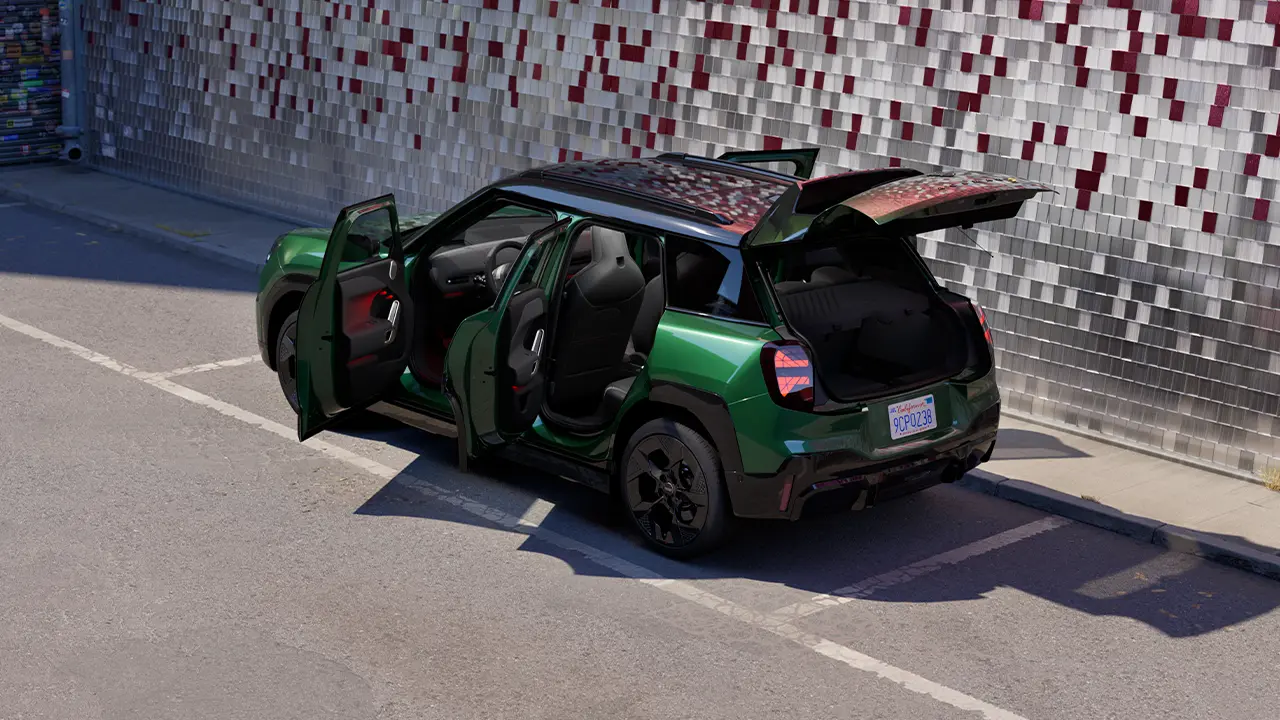 Rear side view of parking all-electric MINI Aceman in British Racing Green in parking lot with all doors open and coloured wall in the background