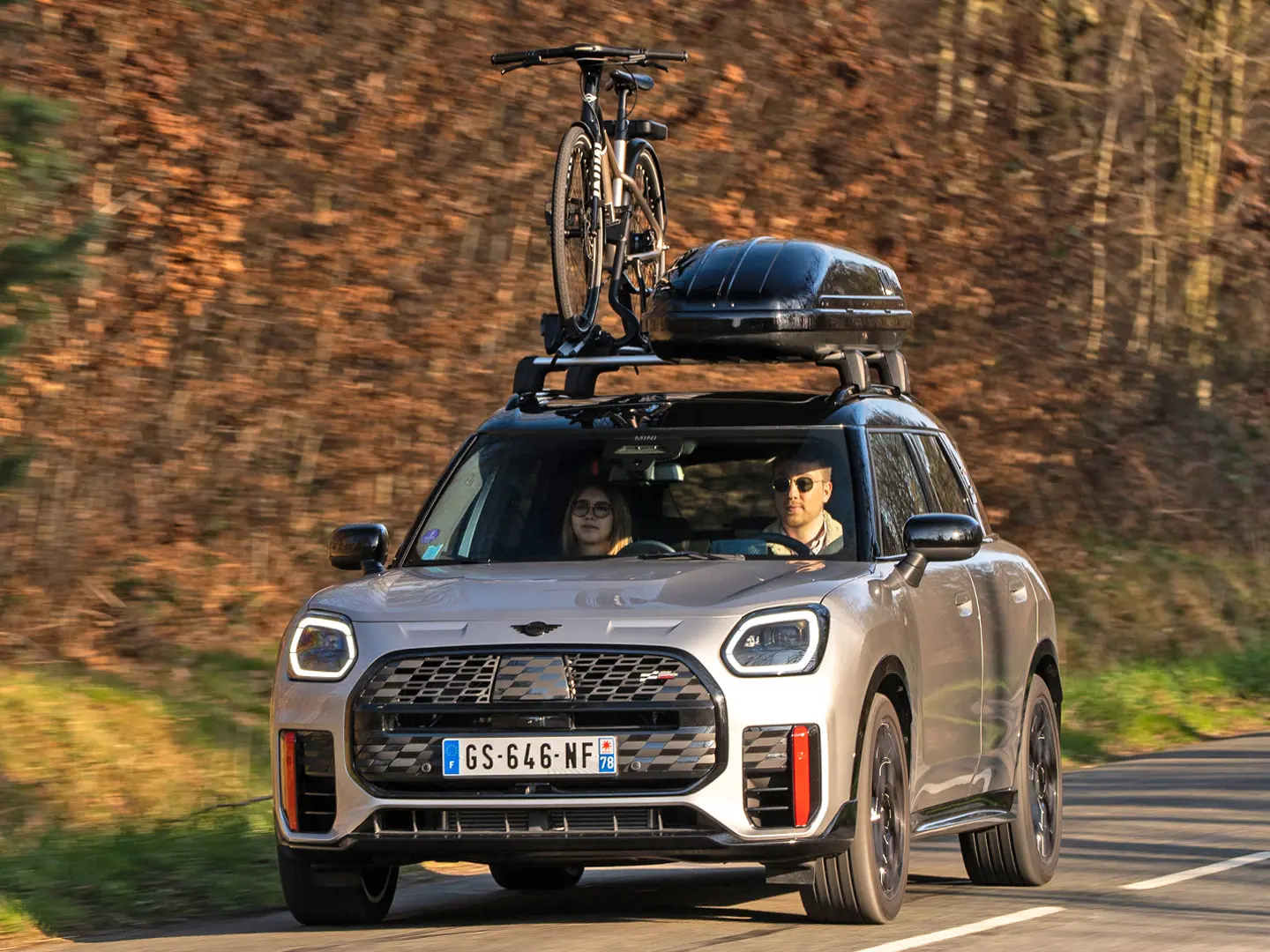 MINI Countryman Cooper（シルバー）が、MINIルーフ・ラックとMINIルーフ・ボックスを装着し、さらに自転車一台を載せたMINIルーフ・サイクル・キャリアを搭載した状態で、山道を走行しています。フロントビュー（正面）からは、MINI John Cooper Works ウィングレットも確認できます。 