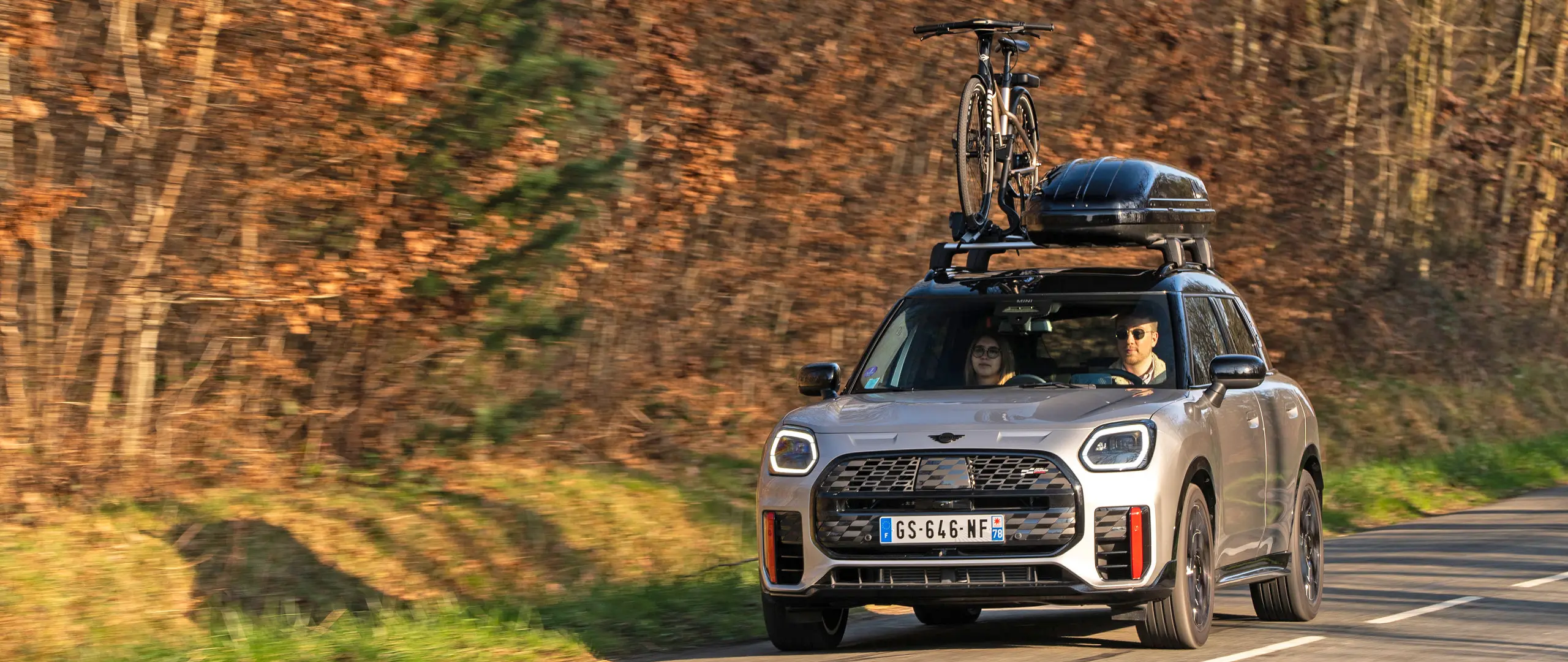 MINI Countryman Cooper（シルバー）が、MINIルーフ・ラックとMINIルーフ・ボックスを装着し、さらに自転車一台を載せたMINIルーフ・サイクル・キャリアを搭載した状態で、山道を走行しています。フロントビュー（正面）からは、MINI John Cooper Works ウィングレットも確認できます。 