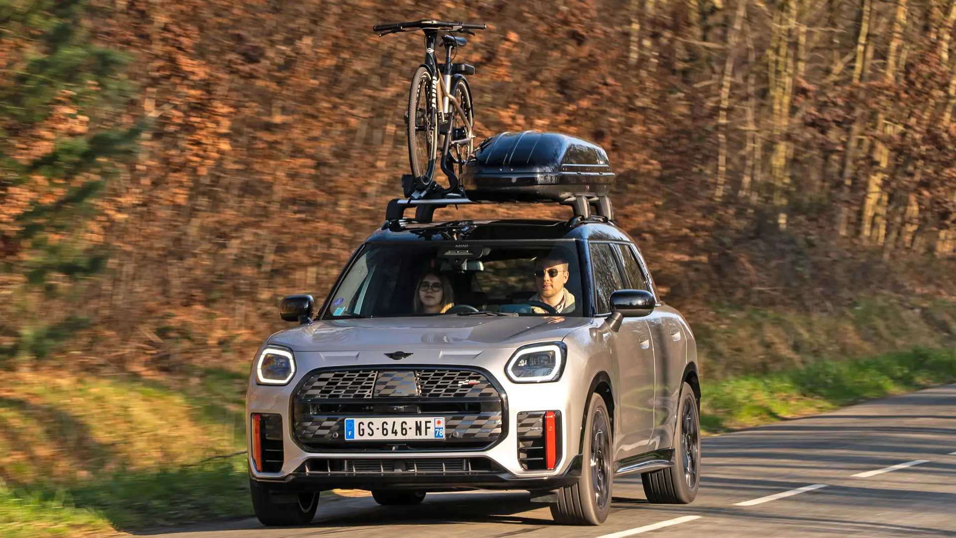 MINI Countryman Cooper（シルバー）が、MINIルーフ・ラックとMINIルーフ・ボックスを装着し、さらに自転車一台を載せたMINIルーフ・サイクル・キャリアを搭載した状態で、山道を走行しています。フロントビュー（正面）からは、MINI John Cooper Works ウィングレットも確認できます。 