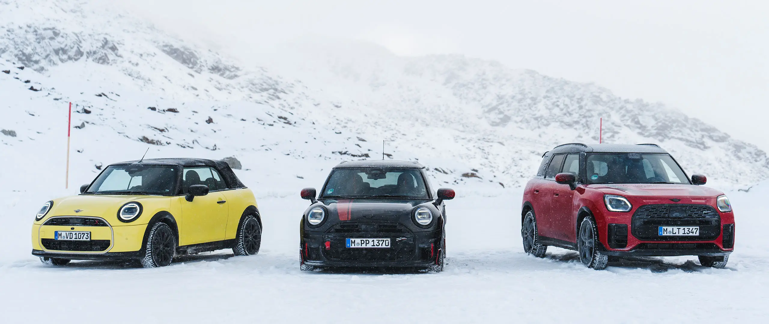 雪に覆われた山道に停車している3台のMINI（正面からの眺め）。
車種とカラーは、MINI Cooper Convertible（サニーサイド・イエロー）、MINI John Cooper Works（ミッドナイト・ブラック II）、MINI Countryman Cooper（チリ・レッド）です。
