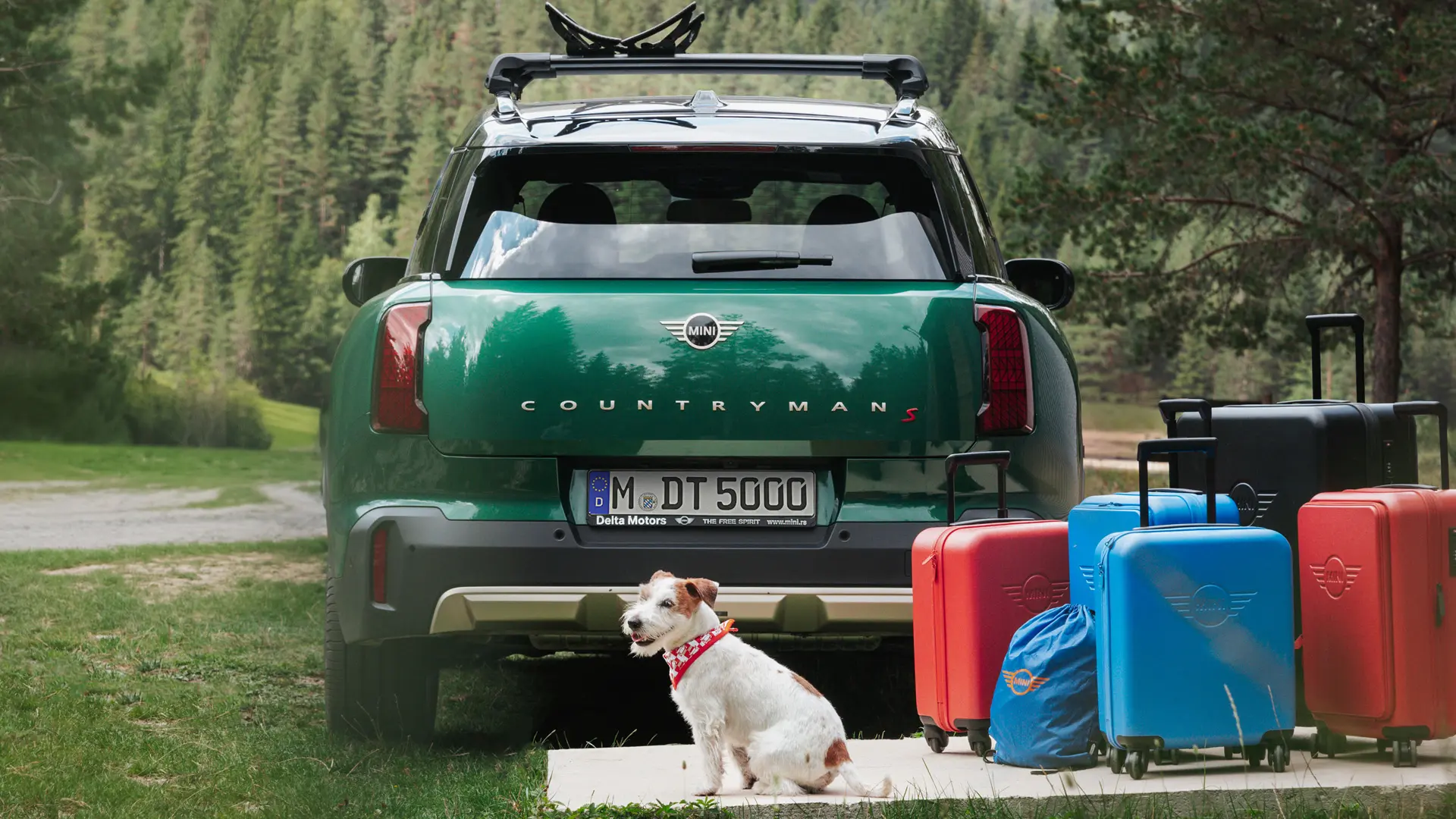 緑の服を着たミニカントリーマンが、ジャック・ラッセルの犬と一緒に車の後ろに立って、MINI Lifestyleストアからさまざまなラゲッジアイテムを購入できます。 