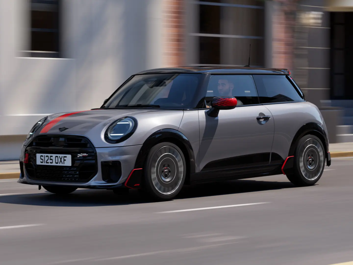 メルティングシルバーのMINI Cooper 3ドアが、チリレッドのジョン・クーパー・ワークス仕様のミラーキャップとボンネットデカール、そしてブラックのルーフスポイラーとウィングレットを装着し、街中を斜め前から捉えたカット