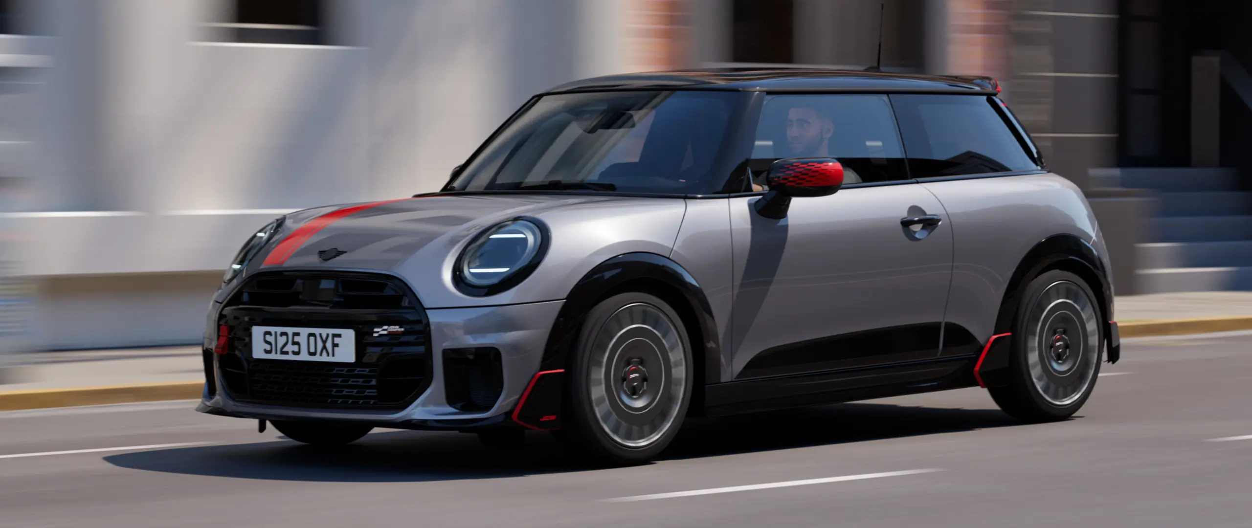 メルティングシルバーのMINI Cooper 3ドアが、チリレッドのジョン・クーパー・ワークス仕様のミラーキャップとボンネットデカール、そしてブラックのルーフスポイラーとウィングレットを装着し、街中を斜め前から捉えたカット