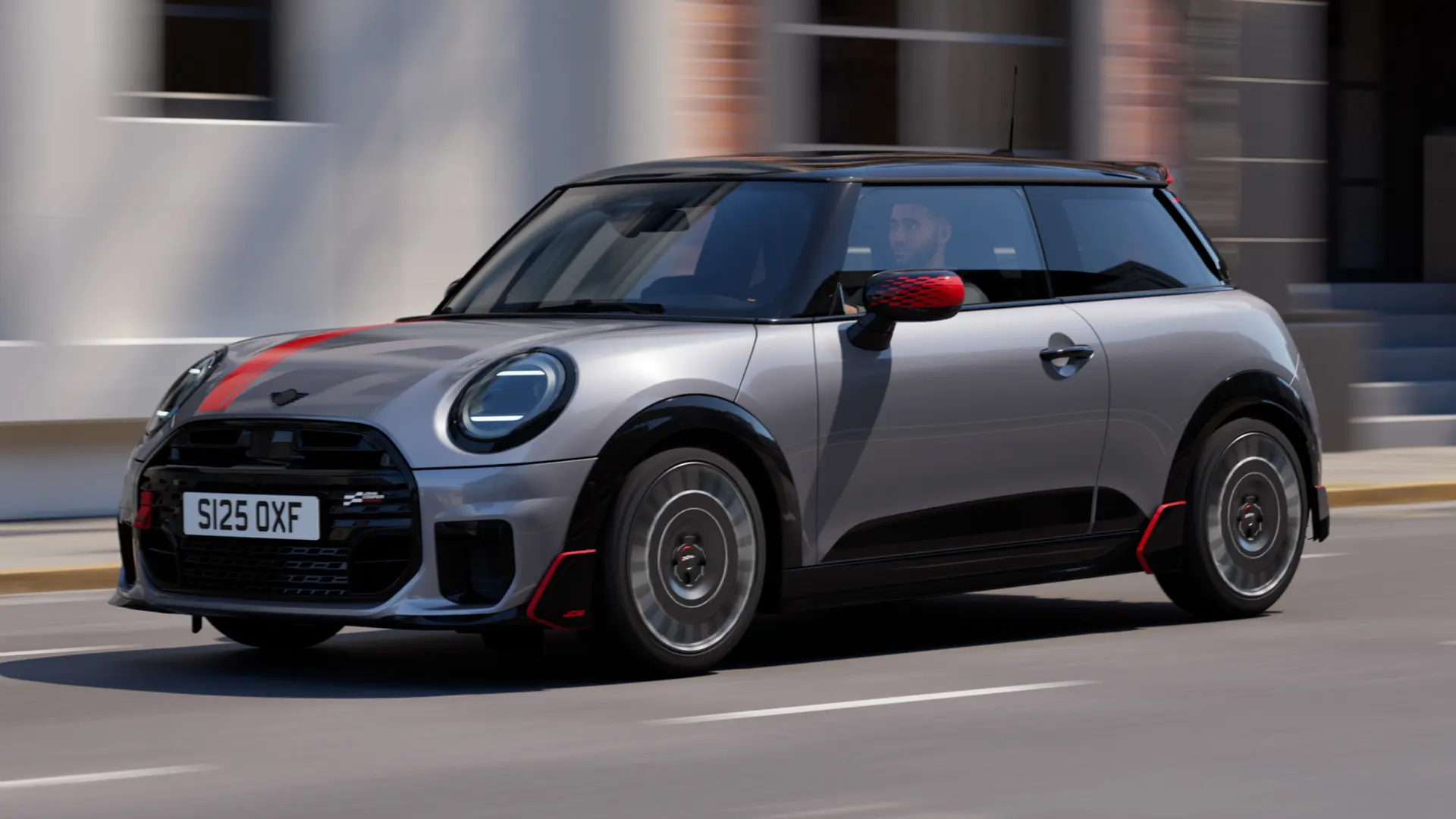 メルティングシルバーのMINI Cooper 3ドアが、チリレッドのジョン・クーパー・ワークス仕様のミラーキャップとボンネットデカール、そしてブラックのルーフスポイラーとウィングレットを装着し、街中を斜め前から捉えたカット